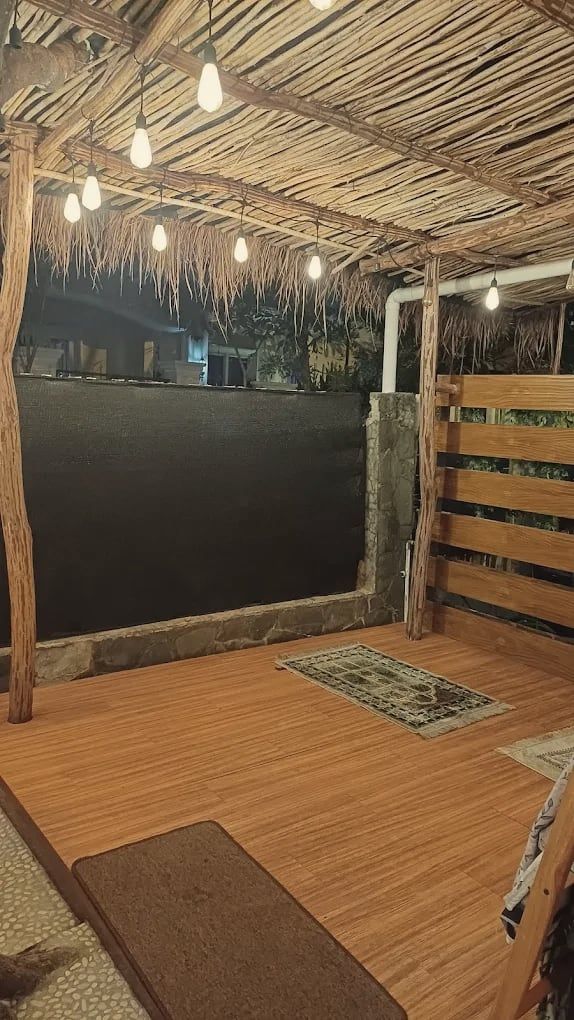 Mushalla terbuka berlantai kayu dengan sajadah di bawah atap rumbia dan lampu bohlam